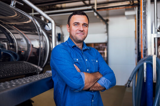 A young smiling man in a denim shirt at the cheese factory. An attractive worker near a milk and a butter making machine. An engineer for the maintenance of a cheese production line. - Powered by Adobe