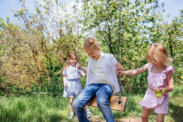 Fototapeta premium Pretty little sisters in summer is rocking brother on swing.