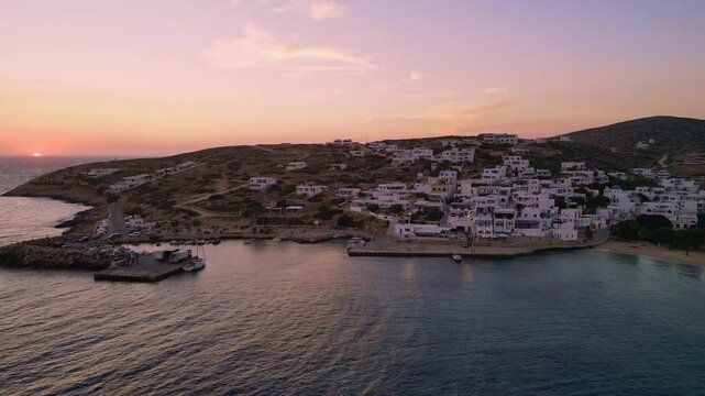 A picturesque aerial view of Stavros Bay on Donousa Island, Greece, with the sun setting over the horizon, casting a warm glow on the white-washed buildings and the calm waters of the Aegean Sea.