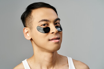 Handsome Asian man in a grey studio using eye patches for his beauty and skincare routine.