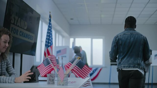 African American man takes ballot for voting from female polling officer at registration table and goes to voting booth. Multiethnic US citizens vote at polling station. United States Election Day.