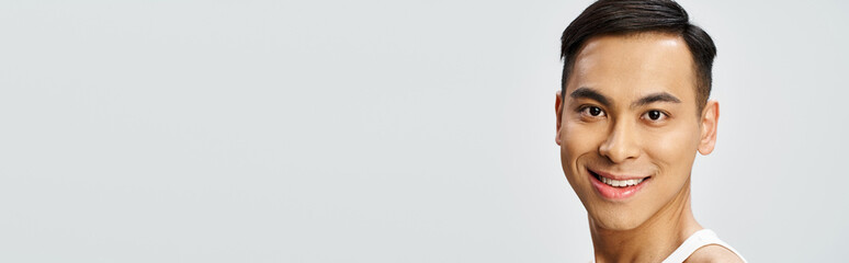 Obraz premium A handsome Asian man in a white tank top smiles serenely at the camera in a grey studio setting.