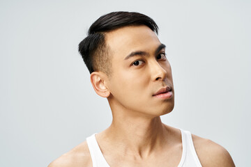 A handsome Asian man in a tank top gazes thoughtfully off to the side in a grey studio setting.