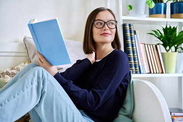 Young woman reading book, sitting on couch at home, engrossed in fiction