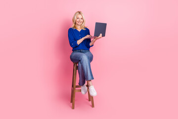 Full body photo of attractive retired woman sit chair hold netbook dressed stylish blue clothes isolated on pink color background