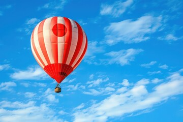 Fototapeta premium A red and white striped hot air balloon, adorned with the Japanese flag design, drifts serenely through a bright blue sky dotted with fluffy white clouds.