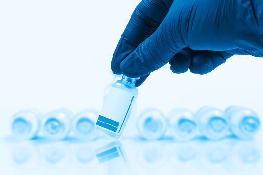 Blue gloved doctor hand holding a vaccine vial in front of a blurred row of vaccine vials on a blue background.