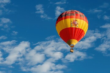Naklejka premium A hot air balloon, adorned with the vibrant colors of the Spanish flag, floats gracefully through a bright blue sky. White clouds drift gently below, adding to the serenity of the scene.