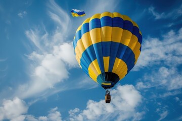 A hot air balloon painted in the colors of the Ukrainian flag ascends gracefully through a vast blue sky dotted with fluffy white clouds.
