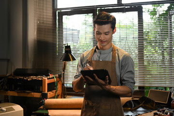 Portrait of handsome craftsman using a digital tablet in leather workshop. Small business and technology concept