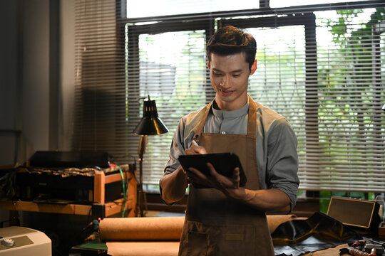 Portrait of handsome craftsman using a digital tablet in leather workshop. Small business and technology concept