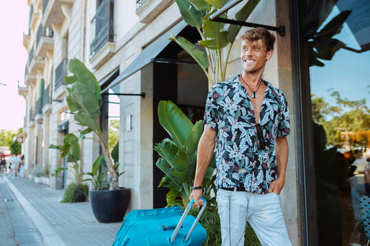 Smiling handsome traveler with suitcase and floral shirt, passing through a chic hotel during summer holidays at city.