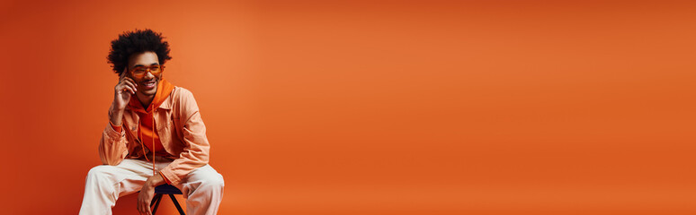 Obraz premium Curly African American man, stylishly dressed, sitting on chair on orange background.