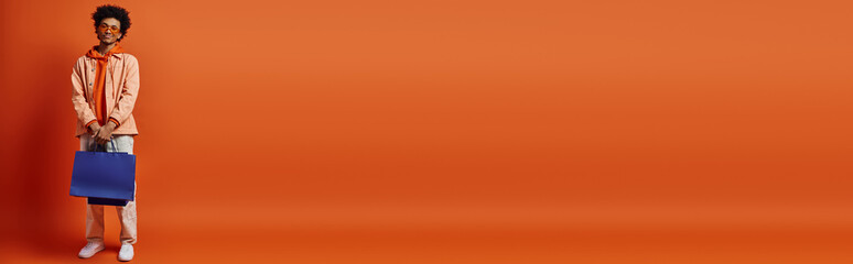 Trendy African American guy in sunglasses stands with blue bag against orange backdrop, exuding emotional energy.