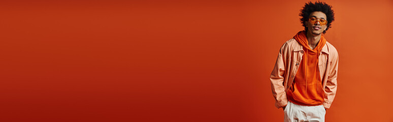 A stylish young African American man, wearing sunglasses and a trendy outfit, poses in front of a bright red wall.