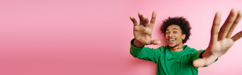 A cheerful young African American man with curly hair in casual wear raising his hands in jubilation on a pink background.