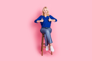 Full body photo of attractive retired woman sit chair thumb up dressed stylish blue clothes isolated on pink color background