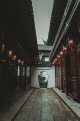 Fototapeta premium A narrow stone path leads through a traditional Chinese garden. The path is lined with dark wooden buildings, some with ornate red doors. A small garden is visible through a white archway, and red lan