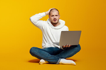 Shocked astonished bearded man in white hoodie looking at camera and holding hand on head while using laptop computer, isolated over yellow background