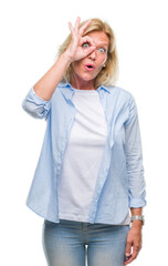 Middle age blonde woman over isolated background doing ok gesture shocked with surprised face, eye looking through fingers. Unbelieving expression.