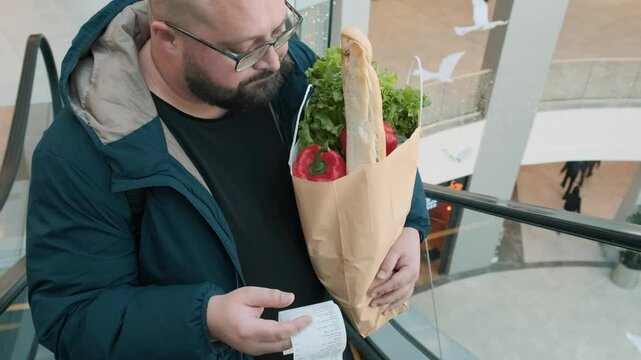 Surprised plus size man with shopping paper bag full of food looks at paper receipt on escalator in supermarket. Male shocked by food inflation and rising prices in shopping mall
