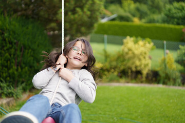 A girl with glasses with curly hair rides on a swing in the backyard in the summer. Happy childhood concept.