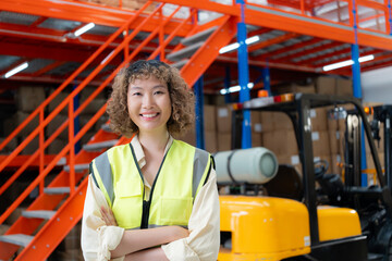 Confident female warehouse worker in a safety vest standing with crossed arms, smiling, with a forklift and shelves in the background.