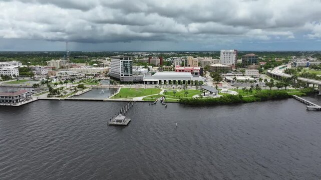 Fort Myers waterfront featuring Centennial Park and yacht basin. Modern buildings and lush green spaces along the Caloosahatchee River. Cityscape with pedestrian walkways and a scenic marina. Aerial.