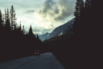 Moody view of a car driving down the road in Canada with the Rocky mountains and clouds in the background. © ovinomis