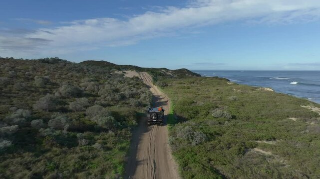 Tracking aerial shot from behind of a 4wd driving along a coastal path, swinging around to the left hand side of the car as it ascends a hill and then swinging around the front to track it backwards.