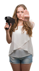 Young blonde woman holding super 8 video camera with open hand doing stop sign with serious and confident expression, defense gesture