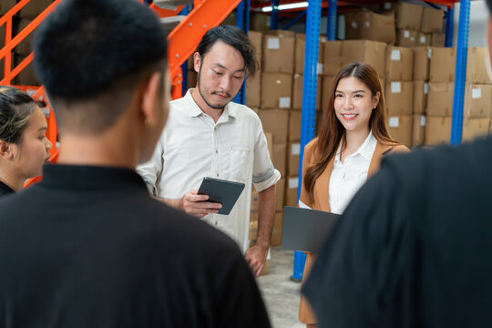 Team gathers for a meeting in a warehouse, with two leaders holding tablets, surrounded by industrial shelving and boxes.
