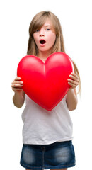 Young blonde toddler holding a red heart scared in shock with a surprise face, afraid and excited with fear expression