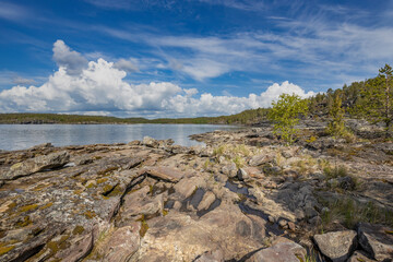 A rocky shoreline stretches out towards a calm lake, the water reflecting the bright blue sky with fluffy white clouds. Lush green trees line the far shore, adding a touch of vibrancy to the scene.