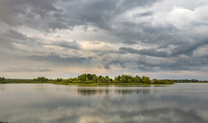 A tranquil scene of a small, forested island in a still lake, framed by a vast expanse of dramatic, grey clouds in the summer sky.