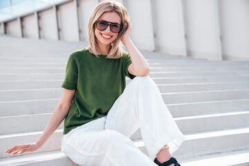 Young beautiful smiling modern woman in trendy summer clothes. Sexy carefree model posing on the street background in sunny day. Positive blond female. Cheerful and happy. Sits at stairs in sunglasses