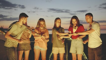 A group of friends is getting ready to roast sausages over a campfire, skewering them under a colorful sunset. Tents are pitched nearby as they enjoy a peaceful evening in nature.