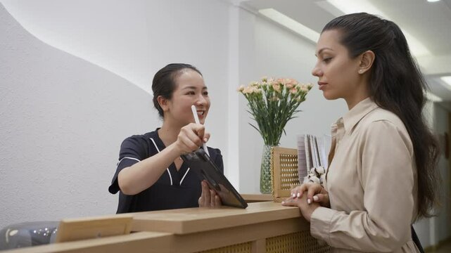Medium shot of cheerful Asian female administrator showing Caucasian woman list of available beauty procedures on digital tablet at spa salon
