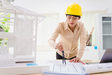 An Asian architect woman sits at her desk, wearing a yellow safety hat. A concrete house structure and toy model illustrate the construction process, showcasing various structural elements.