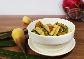 A curry with sliced ​​bamboo shoots, vegetable shoots, chilies, pumpkin in a white porcelain bowl with a saucer, peel, leaves and red fruit next to it. Put it on a white background table.

ไอคอนยืนยัน