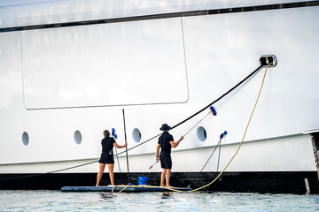 Super yacht crew, two deckhands, male and female cleaning the side of a white mega at dock in...