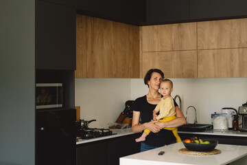 Obraz premium A woman stands in a modern kitchen, holding a baby in her arms. She leans down and kisses the baby on the head. The baby is wearing a yellow onesie and the woman is wearing a black shirt.