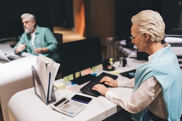 Two professionals working at desks in office. woman types on keyboard, desk equipped with monitor, calculator, documents. man focuses on his laptop. collaborative and productive work environment.