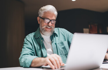 Fototapeta premium Senior man wearing glasses, working a laptop in coworking. Businessman focused and content while analyzing documents. Nighttime office environment, emphasizing concentration and financial dedication