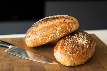 Two loaves of sourdough bread, freshly baked and still warm, are stacked on a wooden surface. The loaves are covered in seeds sesame and sunflower, adding a rustic and flavorful touch.