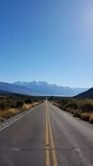 Long straight road leading through a scenic valley with mountain ranges in the distance under a clear blue sky.