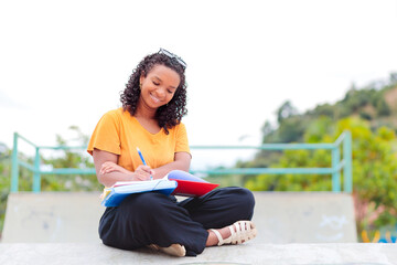 Smiling brazilian Black student with curly hair, wearing a yellow shirt, sits outdoors on campus and writes in a notebook. Green scenery in the background. Casual and relaxed setting