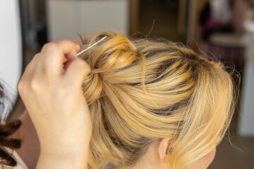 Female stylist styles the hair of blonde woman. Styling hairstyles for wedding or event. High bun of curly hair