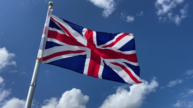 Union Jack British flag flying against a bright blue sky with a few white clouds, on a windy day