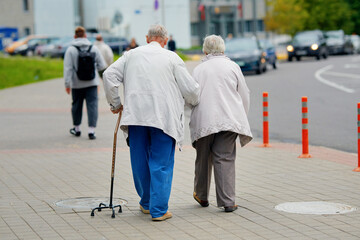 Senior woman supporting sick senior man with walking cane while strolling down the street. Elderly couple supporting each other, enjoying retirement together. Back view of an elderly couple walking